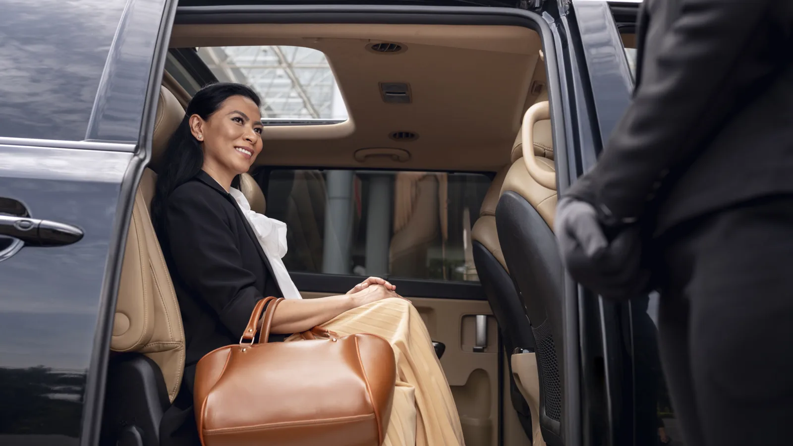 A chauffeur holding the rear door of a black SUV for a passenger with her bag